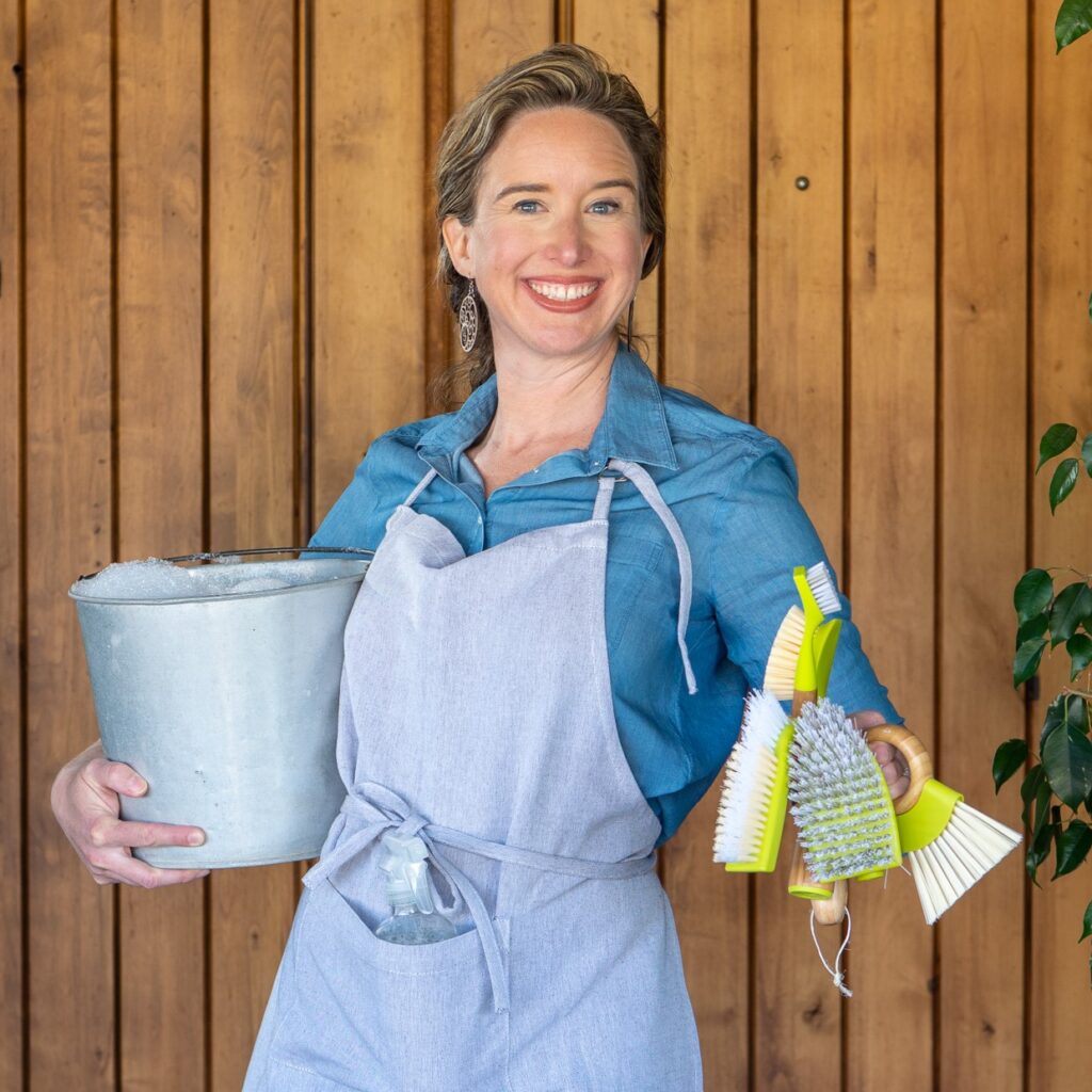 Lisa holding a metal pail in one hand and green cleaning tools in the other.