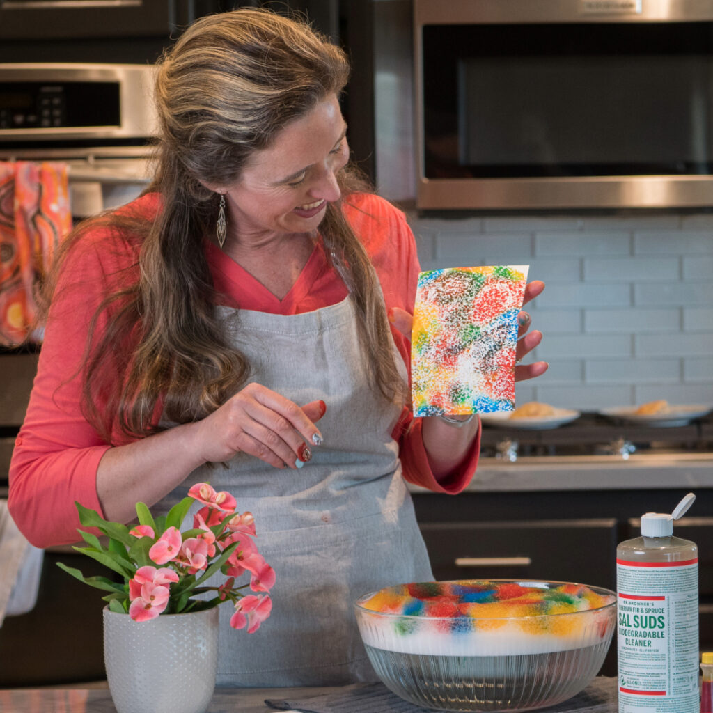 Lisa Bronner holding a colorful sheet of paper in her kitchen with a bowl full of Dr. Bronner's colorful Sal Suds bubbles.