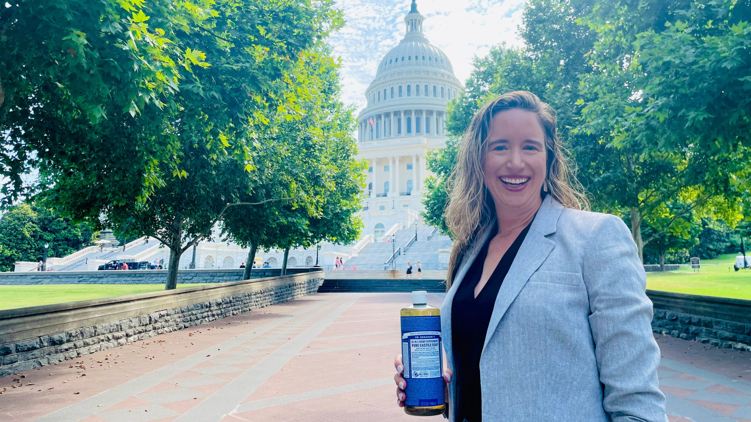 Lisa Bronner standing in front of the capitol building, smiling with a bottle of Dr. Bronner's Castile Soap in her hand.