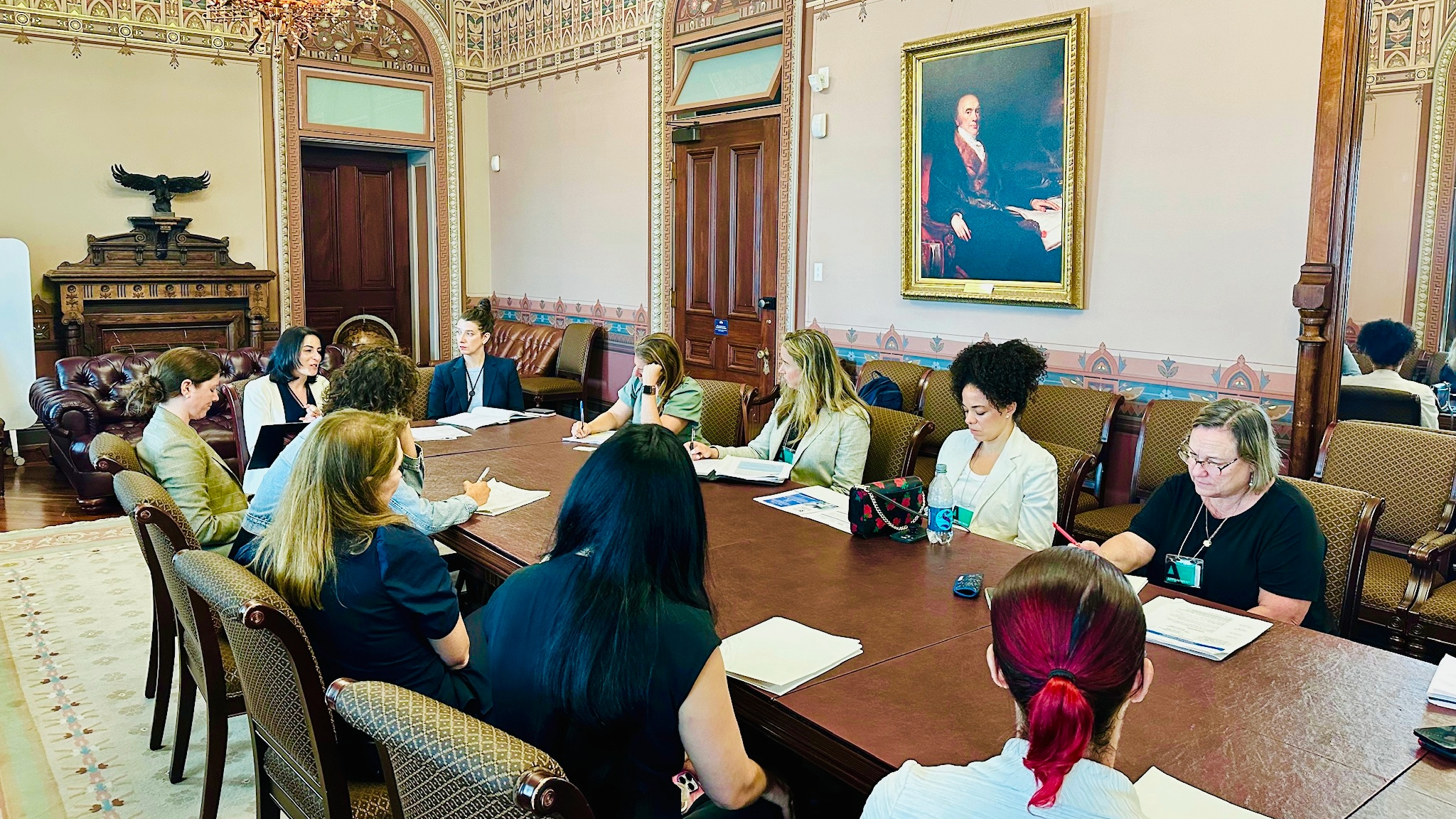 Lisa Bronner and other various staff members sitting about a long table deep in discussion and taking notes in an ornately decorated room. 