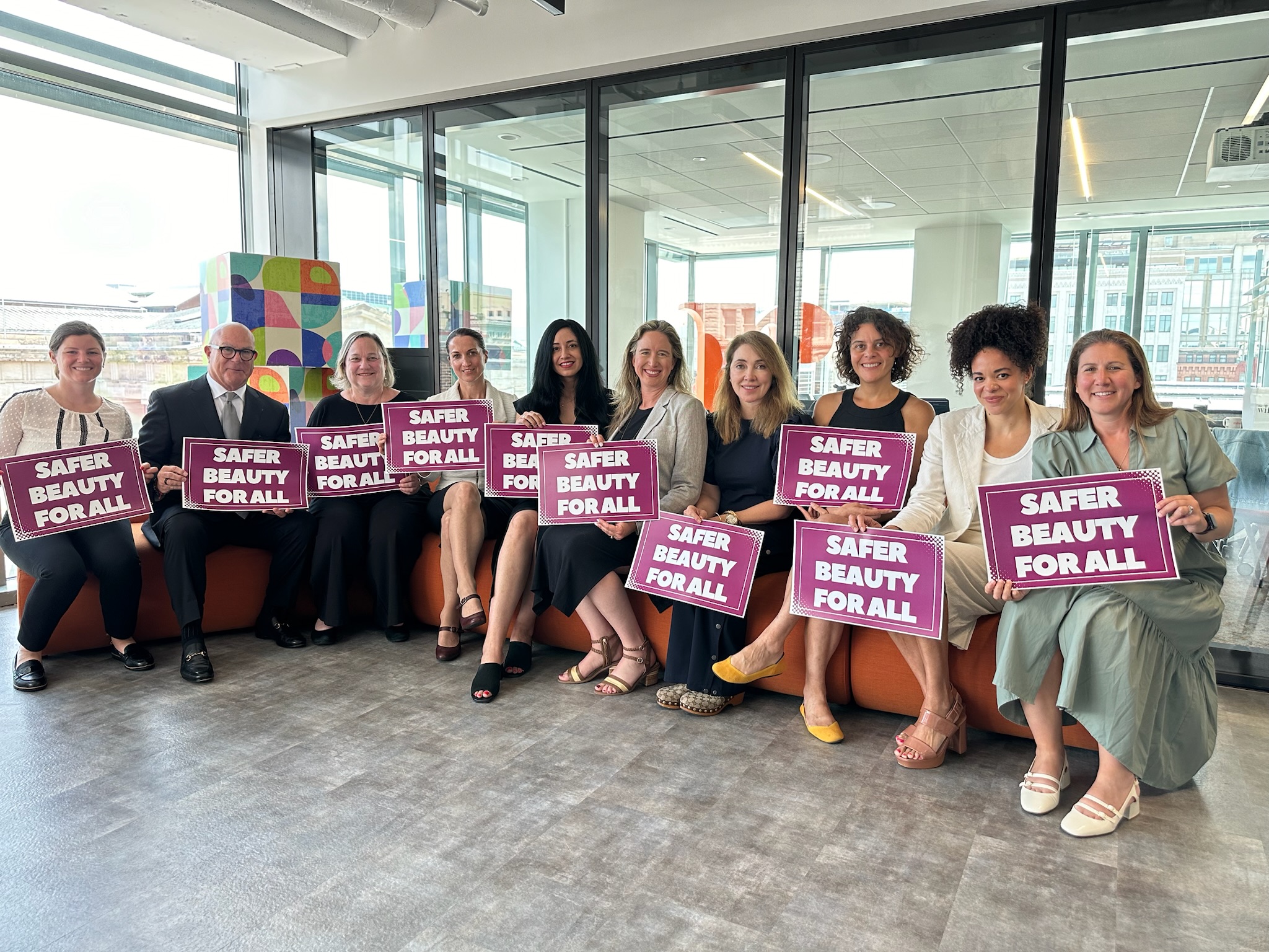 Team of people sitting, smiling at the camera holding signs that say "Safe Beauty For All"