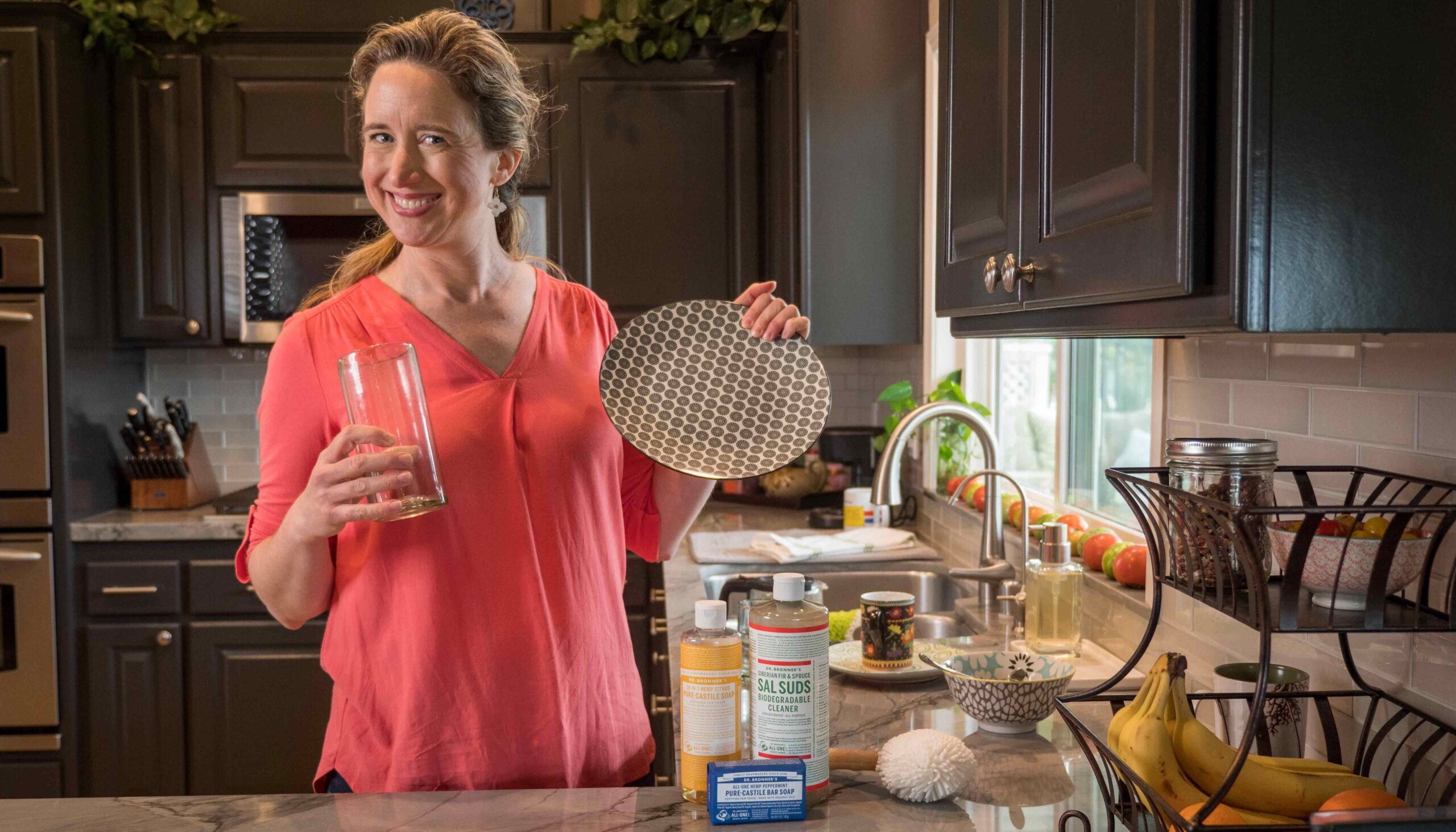 Lisa Bronner, smiling, holding a clean plate and clean glass in front of her sink with Sal Suds and Castile Soap on the counter.