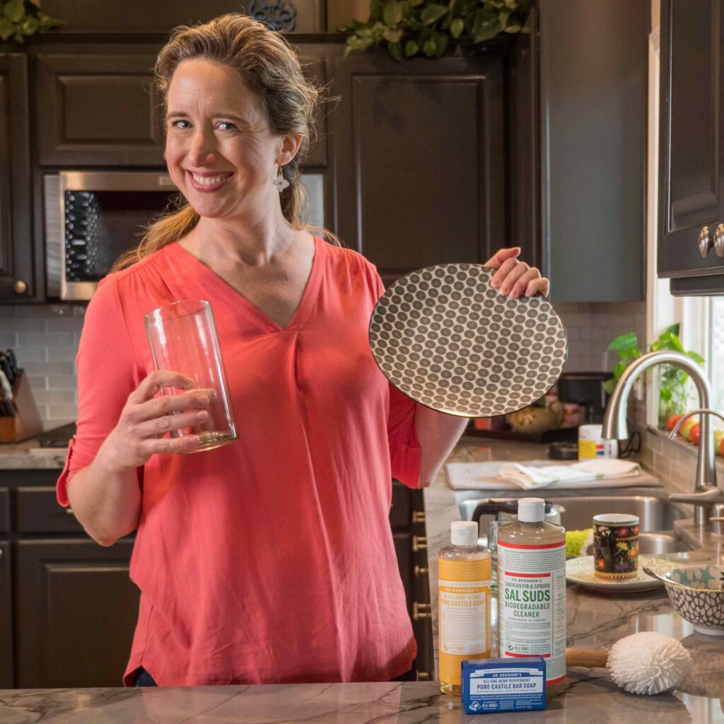 Lisa Bronner, smiling, holding a clean plate and glass in her kitchen with Sal Suds and Castile Soap on the counter.