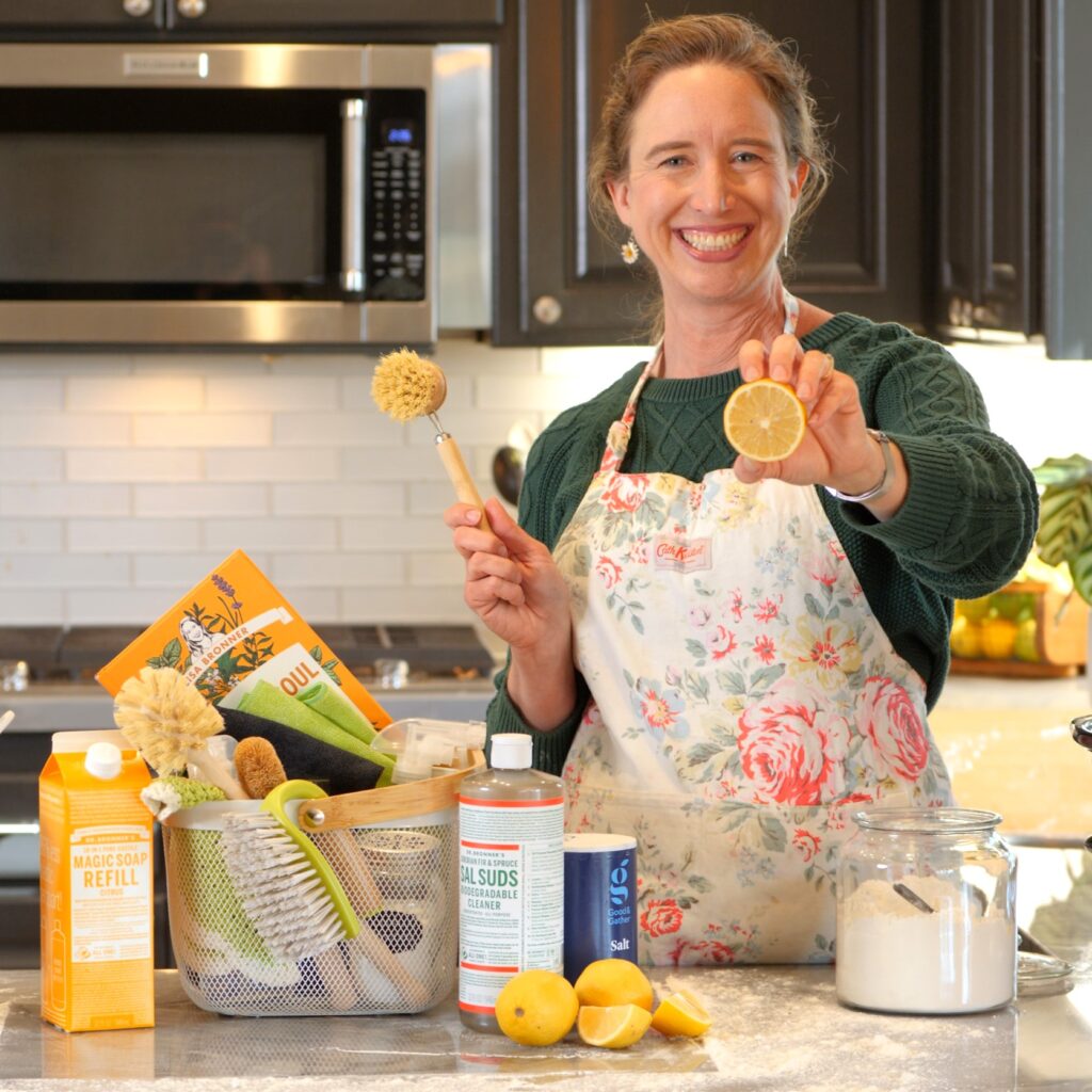 Lisa smiling, holding a lemon and surrounded by cleaning supplies in her kitchen.